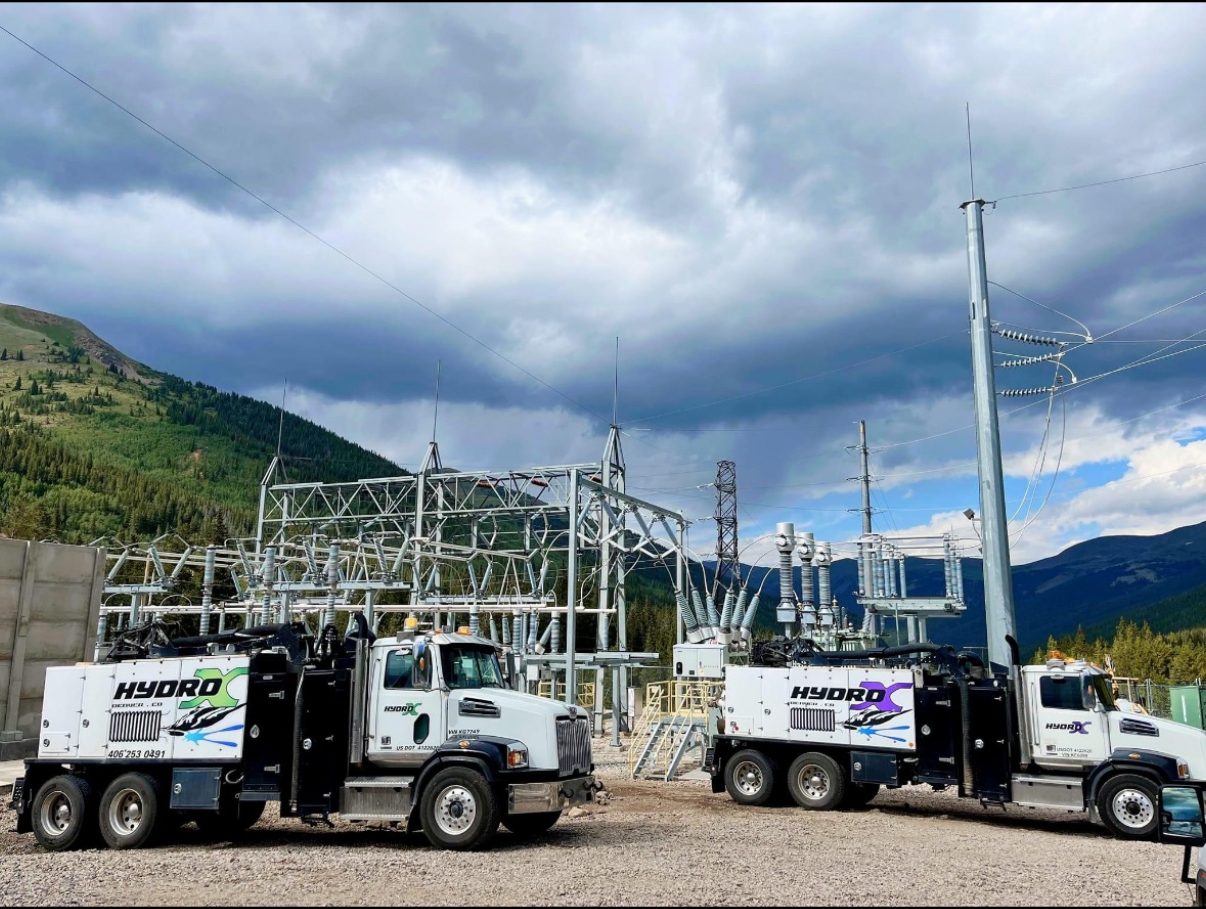 Two white HYDRO-X hydrovac trucks working at an electrical substation in the mountains